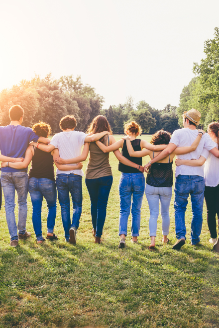 Gruppe Jugendlicher und junger Erwachsener (von hinten fotografiert) auf einer Wiese laufend und sich gegenseitig umarmend