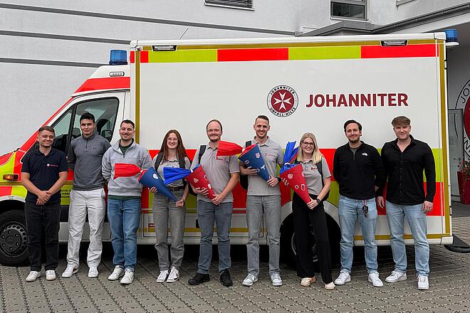 Die neuen Mitarbeitenden im Johanniter-Rettungsdienst sind bereit für ihren Einsatz. Gemeinsam stehen sie in einer Gruppe zusammen und lächeln in die Kamera. Im Hintergrund ist ein Johanniter - Rettungswagen zu sehen.