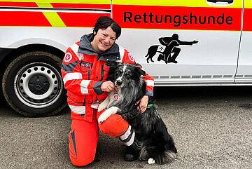 Ein Mitglied der Rettungshundestaffel hockt mit ihrem Hund für ein Foto vor einem Fahrzeug der Staffel.
