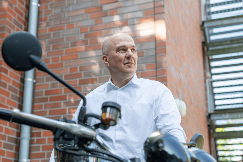 Ein Mann im weißen Hemd steht neben einem Motorrad und hält einen Helm