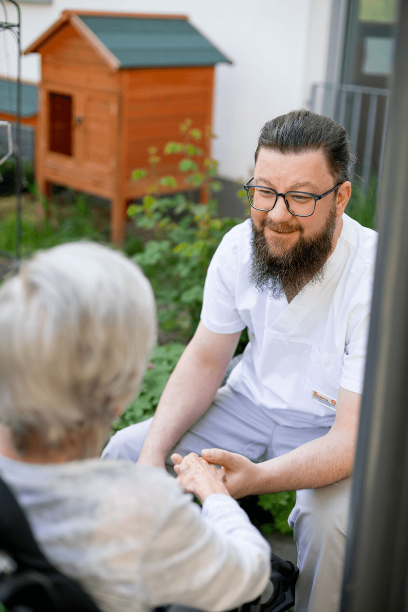 Eine männliche Pflegekraft in weißer Arbeitskleidung kniet und hält die Hände einer Person im Rollstuhl