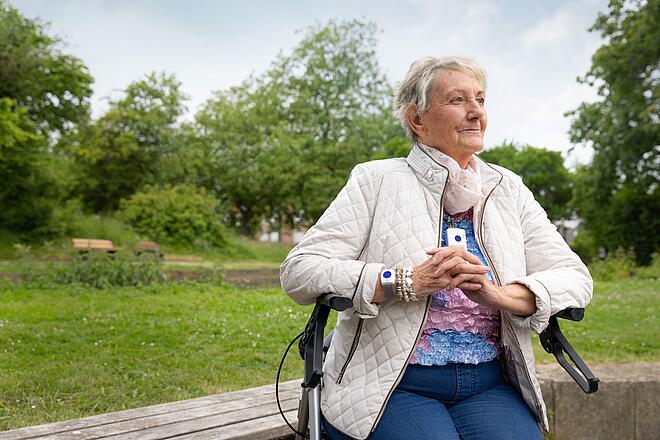 Eine Seniorin in weißer Steppjacke sitzt in einem Park auf einem Rollator. Als Halskette sowie um das Handgelenk trägt sie jeweils ein weißes rechteckiges Gerät mit einem blauen Knopft
