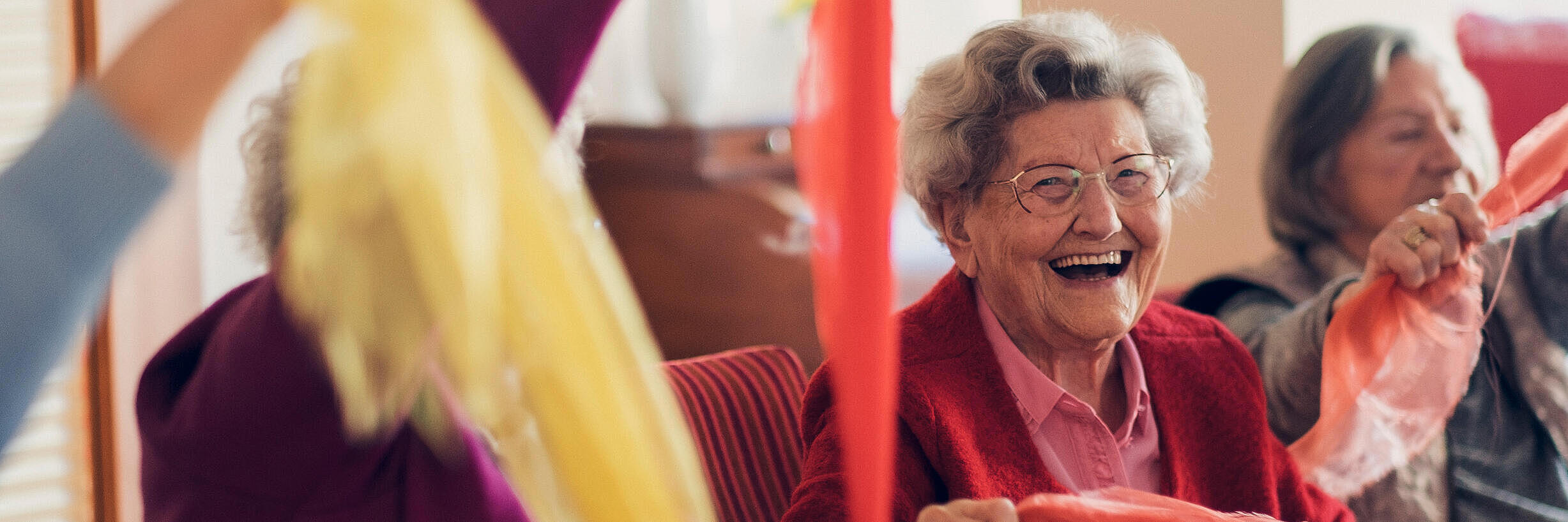 Lachende auf dem Stuhl sitzende Seniorinnen bewegen sich spielend mit einem bunten Tuch