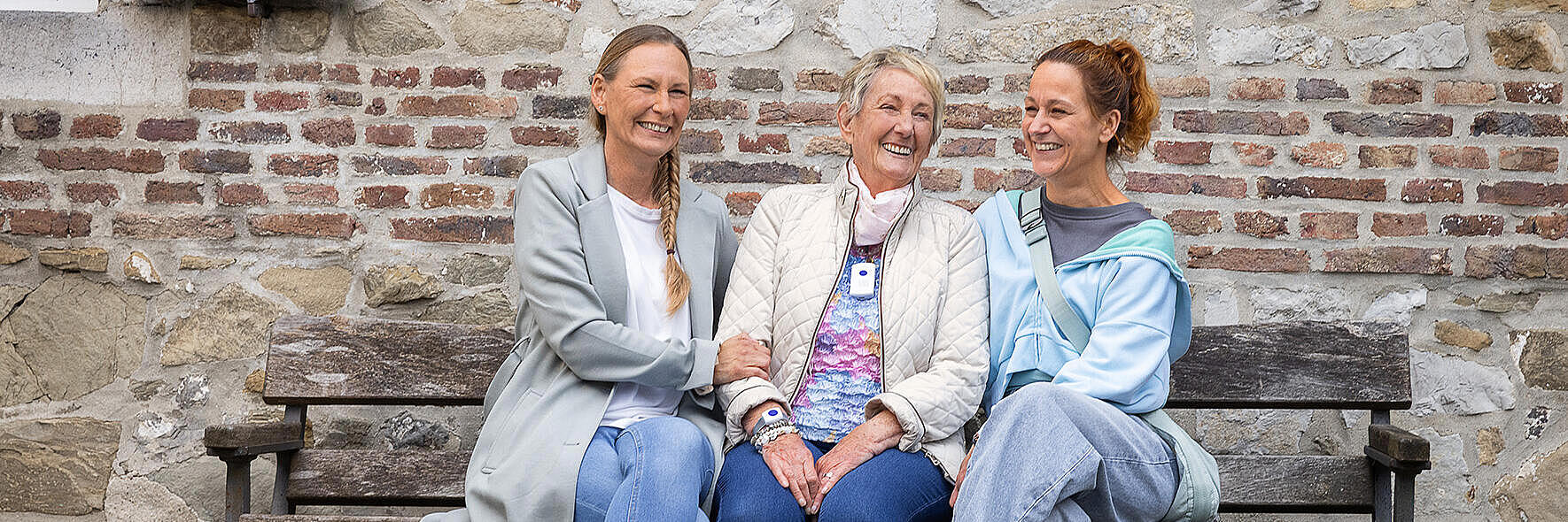 eine ältere Frau sitzt mit zwei jüngeren Frauen auf einer Bank, alle drei lachen