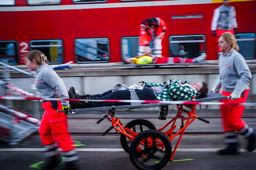 Zwei Johanniter-Rettungskräfte transportieren eine liegende Person auf einer Trage entlang eines Bahnsteigs; Absperrband im Vordergrund und ein stehender Zug im Hintergrund, weitere Verletzte werden versorgt.