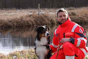 Ein Mitglied der Rettungshundestaffel hockt mit seinem Hund an einem Ufer.