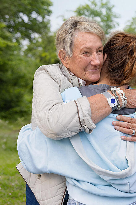 Zwei Frauen umarmen sich. Die ältere Frau trägt einen Handsender am Handgelenk. Im Hintergrund ist ein Park zu sehen.