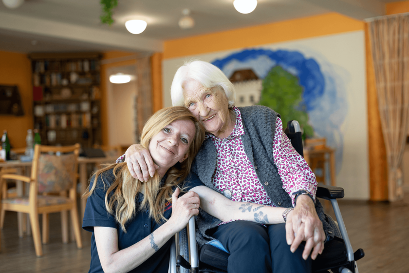 Pflegerin umarmt eine Seniorin im Rollstuhl, beide lächeln herzlich.  